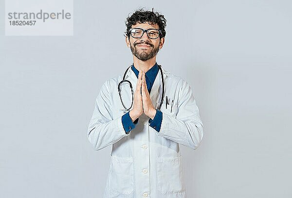 Lächelnder Arzt  der seine Handflächen auf einem isolierten Hintergrund zusammenlegt. Schöner Arzt mit Händen zusammen isoliert. Junger Arzt mit zusammengelegten Handflächen