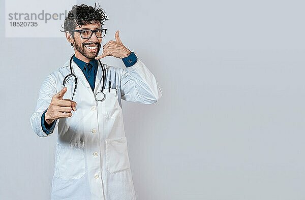 Junger Arzt macht Anruf Geste isoliert. Lächelnder Arzt macht Anruf Geste und zeigt auf die Kamera