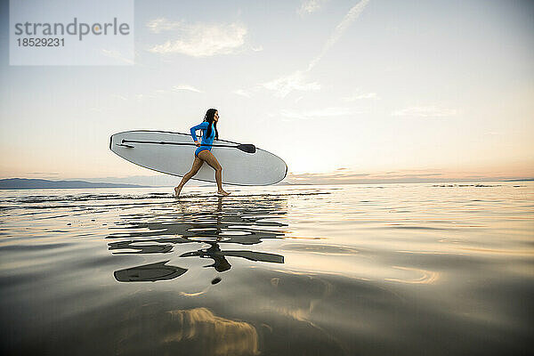 Frau läuft bei Sonnenuntergang mit Paddleboard am Strand