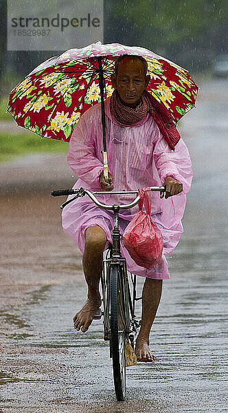 Kambodschanischer Mann mit Regenschirm auf einem Fahrrad im Regen; Siem Reap  Kambodscha