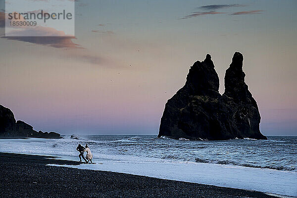 Touristenpaar läuft vor den Wellen  die bei Sonnenuntergang mit der Flut an den Strand kommen; Island