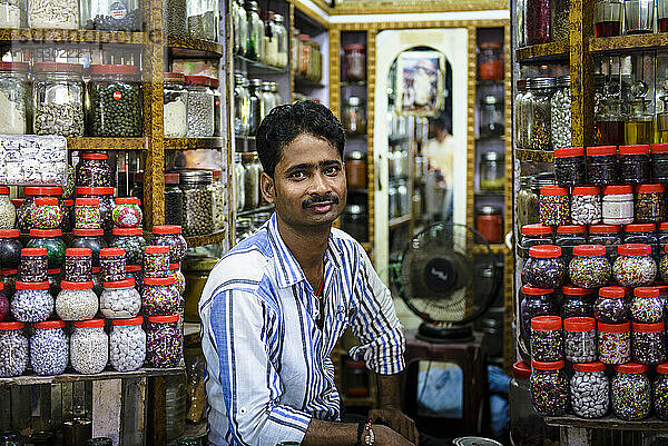 Männlicher Ladenbesitzer sitzt in seinem Süßwarenladen voller Gläser mit Süßigkeiten; Varanasi  Indien