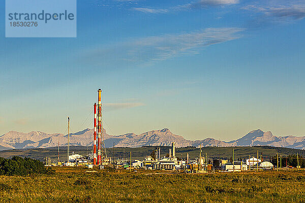 Gaswerk mit rot-weiß gestreiften Türmen  die bei Sonnenaufgang im warmen Licht leuchten  mit Bergkette und blauem Himmel und Wolken im Hintergrund  westlich von Cochrane  Alberta; Alberta  Kanada