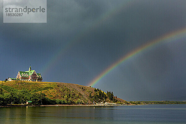 Doppelter Regenbogen in einem dunklen Himmel über einem See mit einem historischen hölzernen Hotelgebäude am Hang über dem See  das bei Sonnenuntergang in warmem Licht erstrahlt; Waterton  Alberta  Kanada