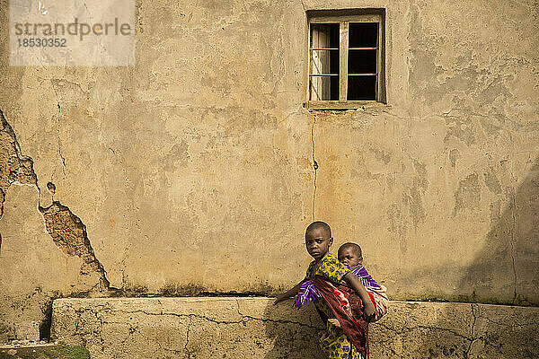 Älteres Kind trägt ein jüngeres auf dem Rücken an einer Mauer; Ruanda