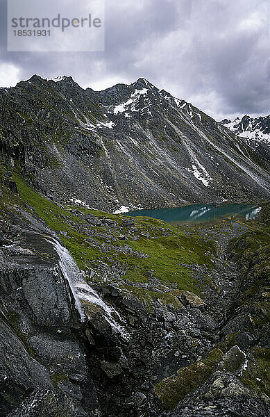 Wasserfall und Granitgipfel am Arch Angle Hatcher Pass unter grauem  bewölktem Himmel mit dem dunkel türkisfarbenen Wasser der Reed Lakes in der Nähe der Independence Mine; Palmer  Alaska  Vereinigte Staaten von Amerika