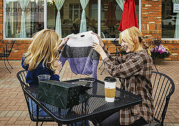 Zwei Teenager genießen die gemeinsame Zeit und sitzen an einem Kaffeetisch in einem Straßencafé  als ein Freund den anderen mit einem Pullover als Geschenk überrascht; St. Albert  Alberta  Kanada