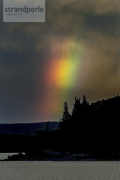 Dramatischer leuchtender Regenbogen über einem See mit leuchtenden Wolken und Silhouette eines Hügels; Waterton  Alberta  Kanada