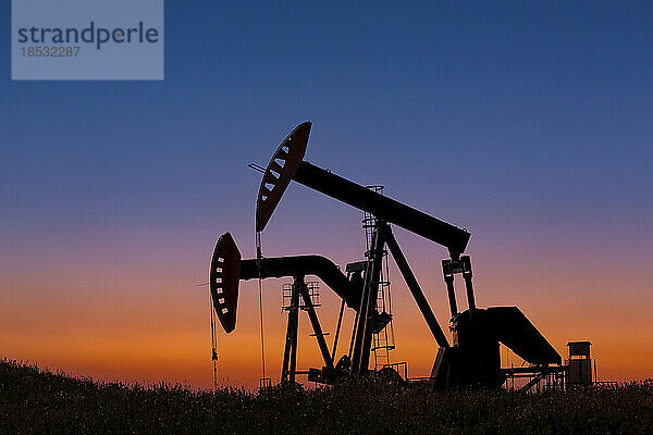 Silhouette von zwei Pumpjacks bei Sonnenaufgang mit leuchtendem Himmel im Hintergrund  westlich von Airdrie; Alberta  Kanada