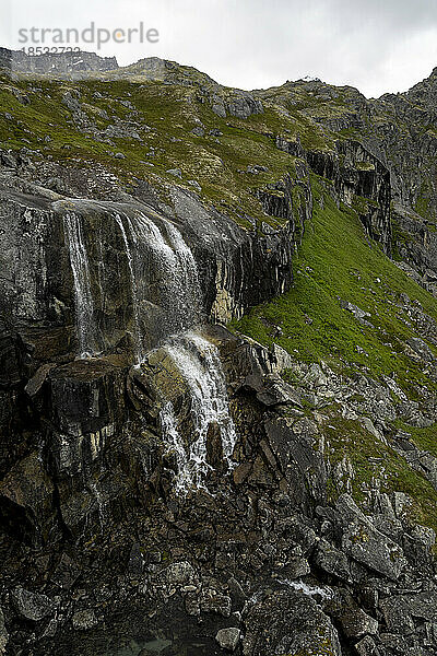 Reed Falls  ein Gletscherwasserfall  der über den grasbewachsenen  felsigen Berghang am Archangel Hatcher Pass unter einem grauen  bewölkten Himmel in der Nähe der Independence Mine fließt; Palmer  Alaska  Vereinigte Staaten von Amerika