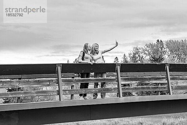 Schwarz-Weiß-Bild von zwei Mädchen im Teenageralter  die auf einer Brücke stehen und ein Selbstporträt mit einem Mobiltelefon aufnehmen  während sie in einem Stadtpark Zeit miteinander verbringen; St. Albert  Alberta  Kanada.