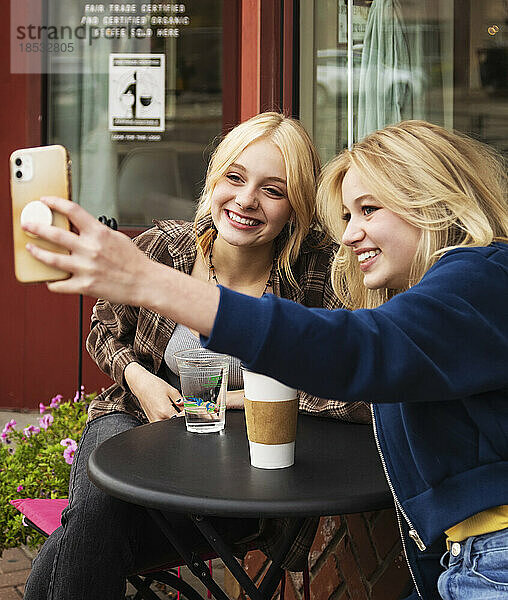 Zwei Teenager genießen die gemeinsame Zeit in einem Café im Freien und verbringen Zeit mit ihren Smartphones  um Selbstporträts zu machen; St. Albert  Alberta  Kanada