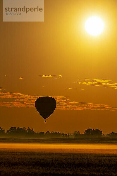 Silhouette eines Heißluftballons in einem orange leuchtenden Himmel bei Sonnenaufgang über einem nebelverhangenen Feld und dem Sonnenaufgang  östlich von Calgary  Alberta; Alberta  Kanada