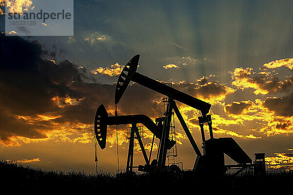 Silhouette von zwei Pumpjacks bei Sonnenaufgang mit warmem  glühendem Himmel und Sonnenstrahlen durch die Wolken im Hintergrund  westlich von Airdrie; Alberta  Kanada