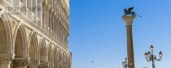 Die geflügelte Löwenstatue auf der Markus-Säule auf dem Markusplatz mit den weißen Bögen des Dogenpalastes (Palazzo Ducale) vor einem blauen Himmel; Venetien  Venedig  Italien