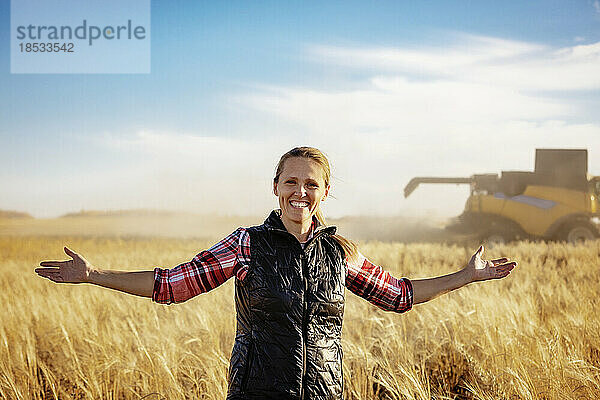 Porträt einer reifen Bäuerin  die mit ausgestreckten Armen in einem Getreidefeld steht und während der Ernte für die Kamera posiert  während im Hintergrund ein Mähdrescher bei Sonnenuntergang arbeitet; Alcomdale  Alberta  Kanada