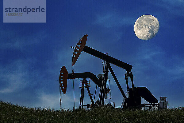 Silhouette von zwei Pumpjacks mit dunkelblauem Himmel und meist vollem Mond im Hintergrund  westlich von Airdrie; Alberta  Kanada