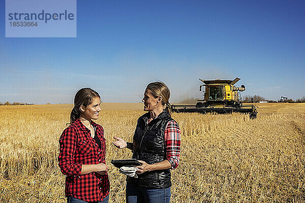 Eine reife Landwirtin steht auf einem Feld und arbeitet zusammen mit einer jungen Frau an der Ernte  wobei sie eine fortschrittliche landwirtschaftliche Software auf einem Pad verwenden  während im Hintergrund ein Mähdrescher arbeitet; Alcomdale  Alberta  Kanada