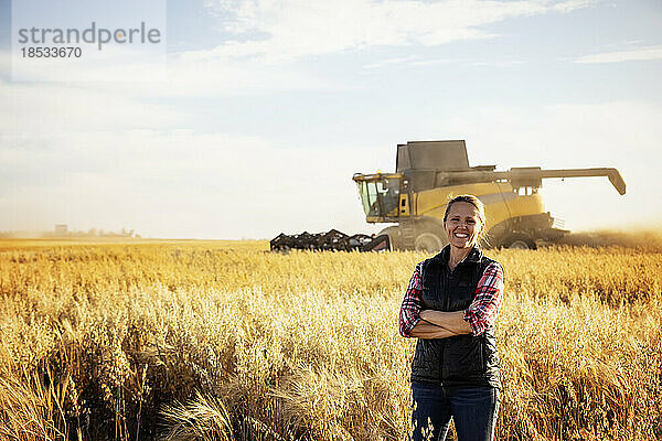 Porträt einer reifen Bäuerin  die während der Ernte in einem Getreidefeld steht und für die Kamera posiert  während im Hintergrund bei Sonnenuntergang ein Mähdrescher arbeitet; Alcomdale  Alberta  Kanada