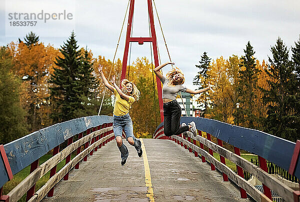 Zwei Mädchen im Teenageralter laufen über eine Brücke und springen in einem Stadtpark an einem warmen Herbstnachmittag in die Luft; St. Albert  Alberta  Kanada.