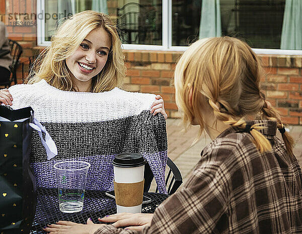 Zwei Teenager genießen die gemeinsame Zeit und sitzen an einem Kaffeetisch in einem Straßencafé  als ein Freund den anderen mit einem Pullover als Geschenk überrascht; St. Albert  Alberta  Kanada