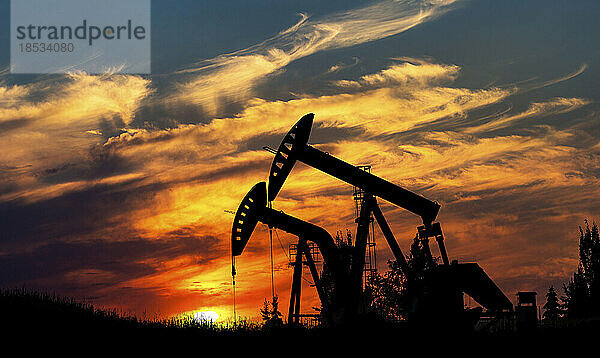 Silhouette von zwei Pumpjacks bei Sonnenaufgang mit dramatischen bunt leuchtenden Wolken im Hintergrund  westlich von Airdrie; Alberta  Kanada