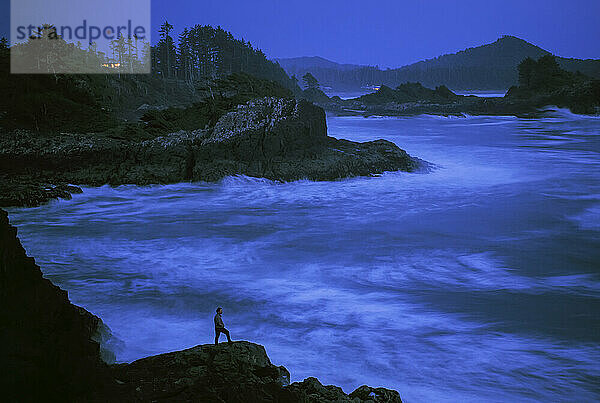 Eine einsame Gestalt beobachtet die Brandung an einer felsigen Küste  während die Nacht hereinbricht; Vancouver Island  British Columbia  Kanada