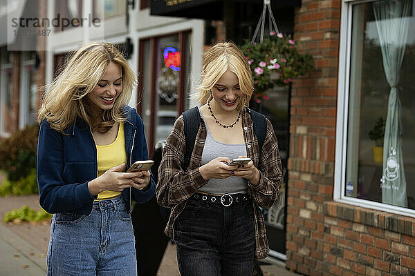 Zwei Teenager genießen einen gemeinsamen Tag beim Einkaufen  gehen eine Straße entlang und benutzen ihre Smartphones an einem Herbsttag; St. Albert  Alberta  Kanada