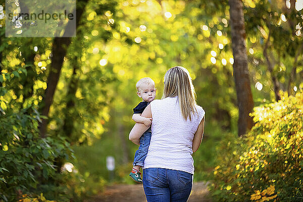 Mutter geht mit ihrem kleinen Jungen im Freien in einem Park spazieren  Blick auf den Rücken der Mutter  während das Baby in die Kamera schaut; Edmonton  Alberta  Kanada