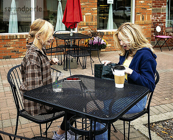 Zwei Teenager genießen die gemeinsame Zeit und sitzen an einem Kaffeetisch in einem Straßencafé  als ein Freund den anderen mit einem Geschenk überrascht; St. Albert  Alberta  Kanada
