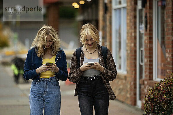 Zwei Teenager genießen einen gemeinsamen Tag beim Einkaufen  gehen eine Straße entlang und benutzen ihre Smartphones an einem Herbsttag; St. Albert  Alberta  Kanada