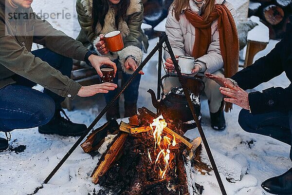 Eine Gruppe von Freunden versammelt sich um ein Lagerfeuer im Hinterhof  trinkt Tee und wärmt sich die Hände. Zwei glückliche Paare  die sich entspannen und die Wintersaison genießen  während sie am Feuer sitzen. Outdoor Winter unterhaltsam