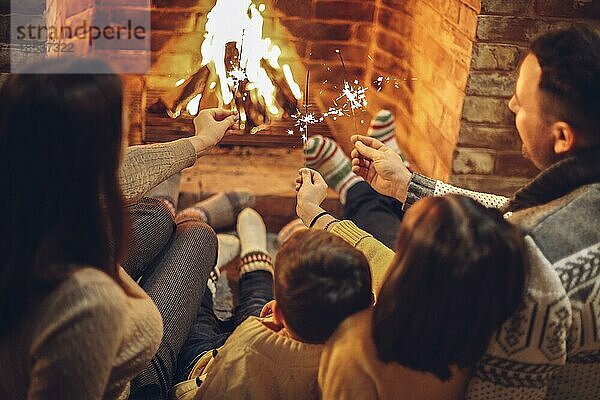 Glückliche junge Familie Eltern mit zwei kleinen Kindern versammeln sich um gemauerten Kamin in gemütlichen und warmen Landhaus während der Weihnachtsferien im Winter. Vater  Mutter und Kinder sitzen am Feuer zu Hause