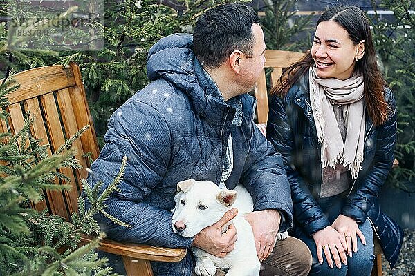 Lovely Paar mit niedlichen Hund sitzen auf Hinterhof außerhalb von jungen Nadelbäumen umgeben  lächelnd süß  während die Kommunikation mit einander auf frischen frostigen Luft  genießen Winterurlaub zusammen