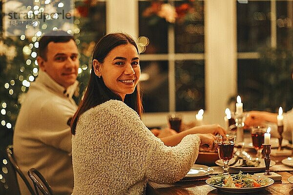 Glückliches junges Familienpaar  das in die Kamera lächelt  während es an einem festlichen Tisch sitzt  der für den Weihnachtsabend gedeckt ist  Mann und Frau  die Silvester in einem gemütlich dekorierten Haus mit einem Weihnachtsbaum im Hintergrund feiern