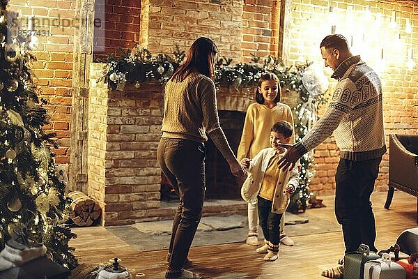 Glückliche Familie Eltern mit zwei Kindern tanzen zusammen in der Nähe von Kamin und Weihnachtsbaum in gemütlichen Landhaus für die Weihnachtsferien dekoriert. Mutter  Vater und Kinder feiern Neujahr im Bauernhaus