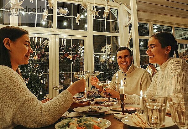 Ein glückliches Familienpaar stößt mit Sektgläsern an  während es Silvester oder Weihnachten mit Freunden feiert und an einem festlich gedeckten Tisch mit traditionellen Speisen und brennenden Kerzen sitzt