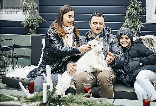 Glückliche Familie mit Hund sitzt auf Bank im Haus Hof während der Winterferien. Liebevolle fröhliche Eltern und Tochter spielen mit Welpe im Hinterhof während der Weihnachtszeit
