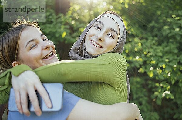 Starke weibliche Freundschaft. Glückliche zwei Teenagermädchen beste Freunde halten die Hände und umarmen  während sie vor einem Park stehen. Multiethnische Freunde