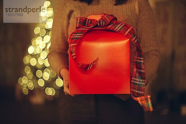 Ausgeschnittenes Foto einer nicht erkennbaren Frau  die eine Weihnachtsgeschenkbox hält  während sie zu Hause mit einem unscharfen Weihnachtsbaum im Hintergrund steht. Selektiver Fokus einer Frau  die ein Neujahrsgeschenk erhält