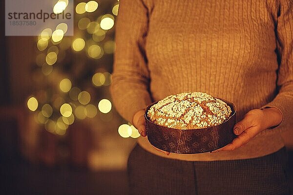 Ausgeschnittene Aufnahme einer Frau  die einen leckeren  frisch gebackenen  hausgemachten Weihnachtskuchen hält  während sie vor einem unscharfen Weihnachtsbaum im Hintergrund steht und Essen für Silvester vorbereitet  selektiver Fokus auf Panettone Kuchen in den Händen
