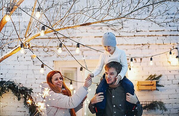 Glückliche Familie mit einem Kind  das vor einem gemauerten Haus steht  das mit Tannenzweigen und schönen Weihnachtslichtern geschmückt ist. Die Eltern und der kleine Junge freuen sich über die Winterferien im Freien