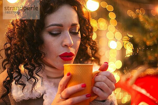 Junge Frau  die ein heißes Getränk aus einem Becher trinkt  während sie vor einem unscharfen  geschmückten Weihnachtsbaum im Freien während der Weihnachtsfeiertage steht  Frau mit festlichem Make up und Frisur  die eine Tasse Tee hält  unscharf