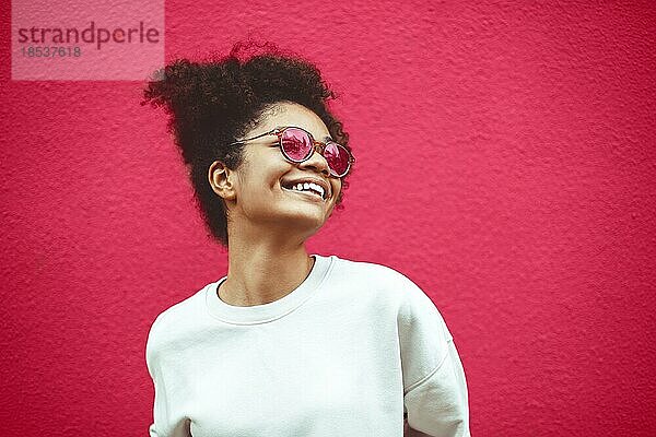 Junge Frau afrikanischer Abstammung mit modischer Sonnenbrille und lockigem Haar  das zu einem hohen Pferdeschwanz gebunden ist. Sie schaut weg und lächelt breit  während sie gerade  perfekte Zähne zeigt und vor einem Wandhintergrund posiert