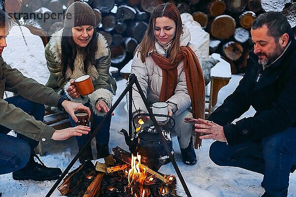 Eine Gruppe von Freunden versammelt sich um ein Lagerfeuer im Hinterhof  trinkt Tee und wärmt sich die Hände. Zwei glückliche Paare  die sich entspannen und die Wintersaison genießen  während sie am Feuer sitzen. Outdoor Winter unterhaltsam