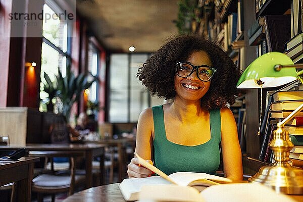 Glückliche afroamerikanische Studentin  die in der Universitätsbibliothek studiert und in die Kamera schaut  mit Brille und grüner Lampe. Lächelndes Teenagermädchen in gemütlicher Bibliothek