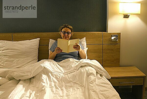 Mittleres Alter kaukasische Frau lesen Buch sitzen auf dem Bett im Schlafzimmer. Menschen  Schlafenszeit und Ruhe Konzept