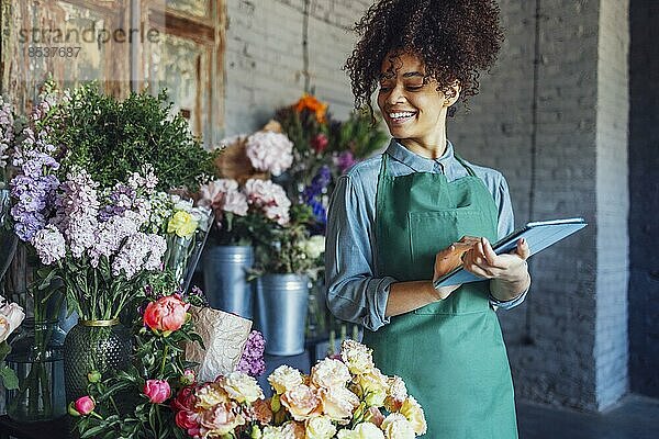 Junge schwarze Frau mit beiger Schürze auf Blumenladen Hintergrund mit Kopie Raum. Porträt einer erfolgreichen afroamerikanischen Frau  die auf die Blumen schaut und einen Tabletcomputer hält