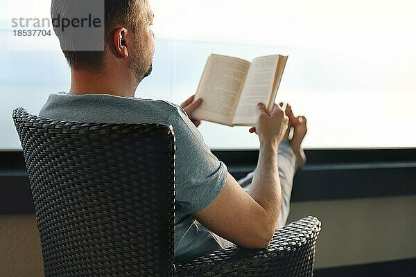 Junger Mann liest Buch in der Nähe von Fenster und suchen Meerblick auf dem Land Gasthof in den Morgen Sonnenaufgang. Solo Reise  Reise  Reise und entspannende Konzept