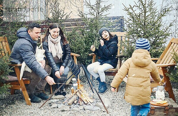 Eine Familie in warmer Kleidung sitzt draußen am brennenden Lagerfeuer  röstet Marshmallows auf Stöcken und unterhält sich  freigestelltes Foto. Angenehmer Zeitvertreib an frischer  frostiger Luft in den Winterferien
